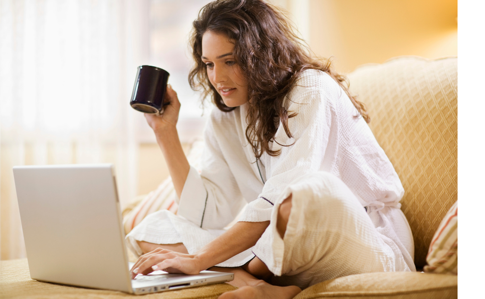 Woman in her own home on the couch looking towards a computer.