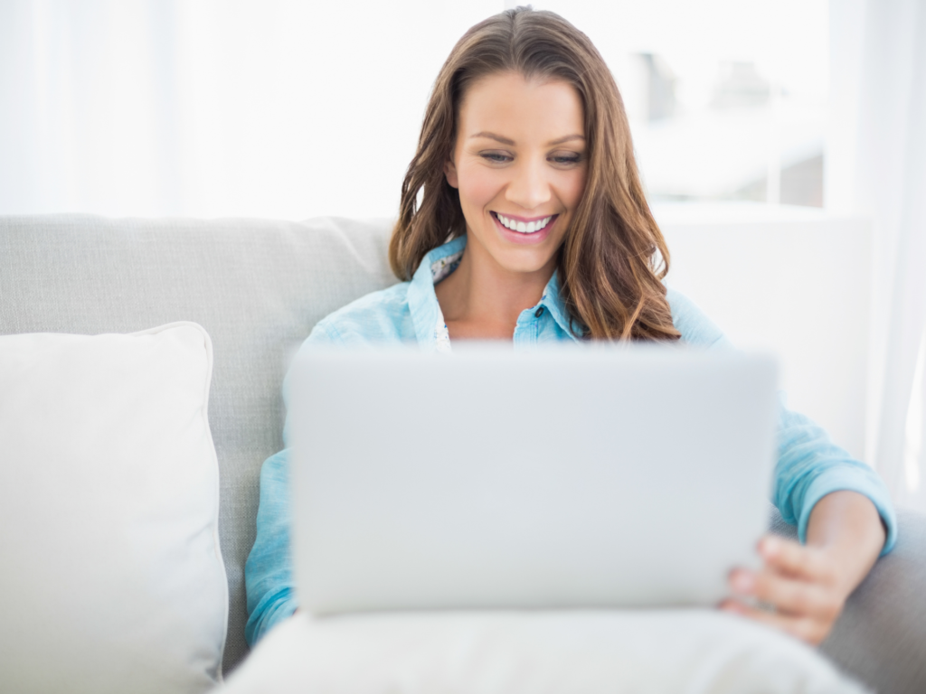 woman smiling at her laptop while she sits on a couch in her home during her online therapy session