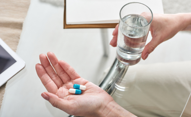  hands holding two pills with a glass of water in the other hand. 