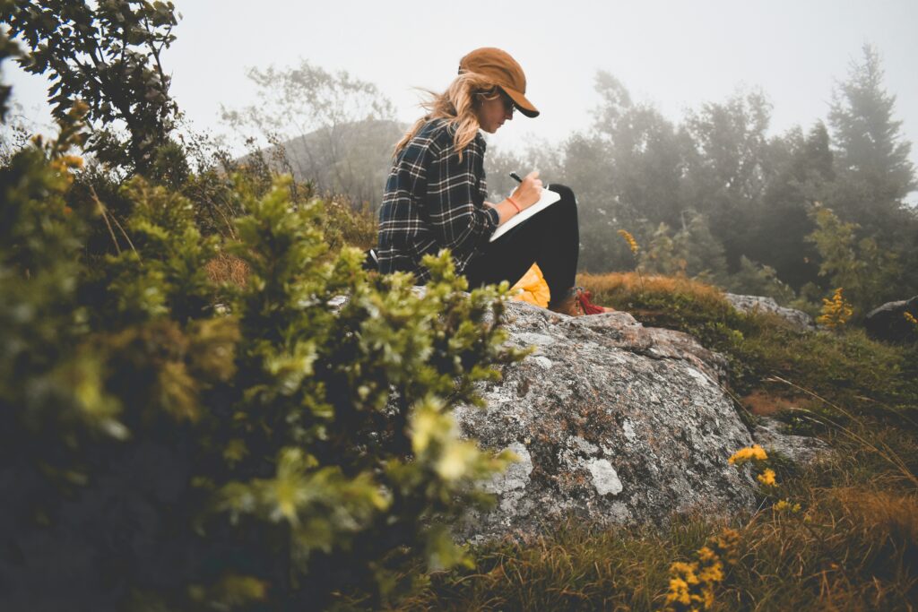 A woman in a plaid shirt and brown cap sits on a mossy rock in a misty, overgrown landscape, journaling outdoors.