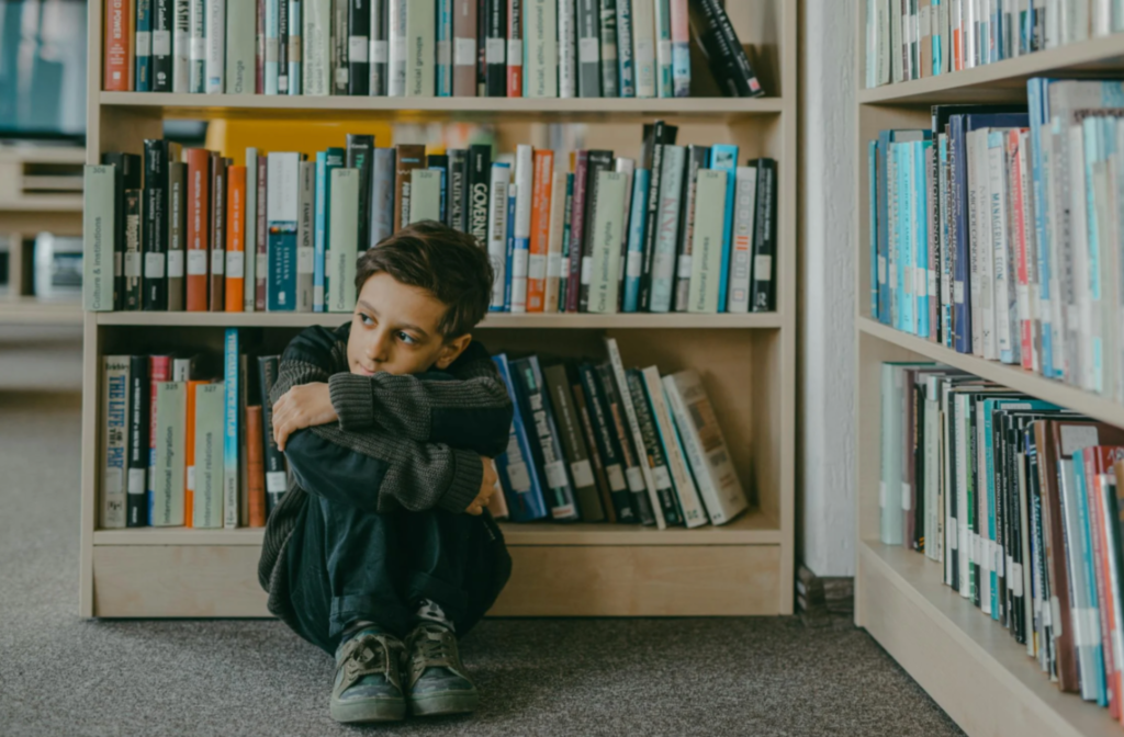 A young boy sitting on the floor between bookshelves in a library, hugging his knees and looking off to the side with a quiet, contemplative expression.