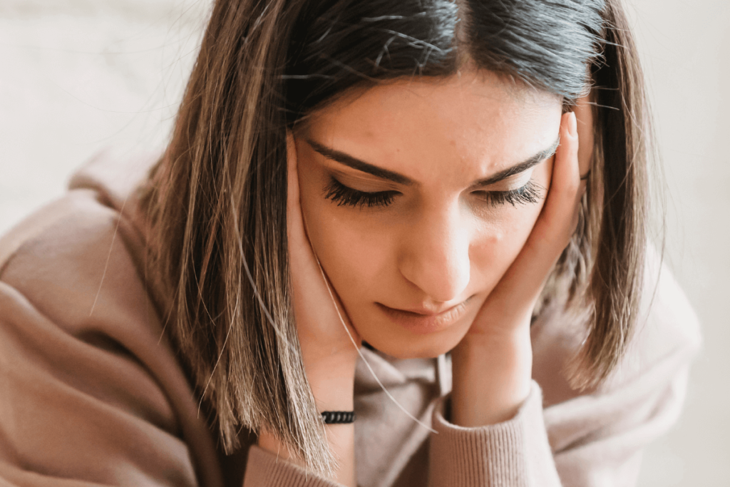 A young woman in a beige hoodie holds her face in her hands, looking down with a worried, distressed expression.