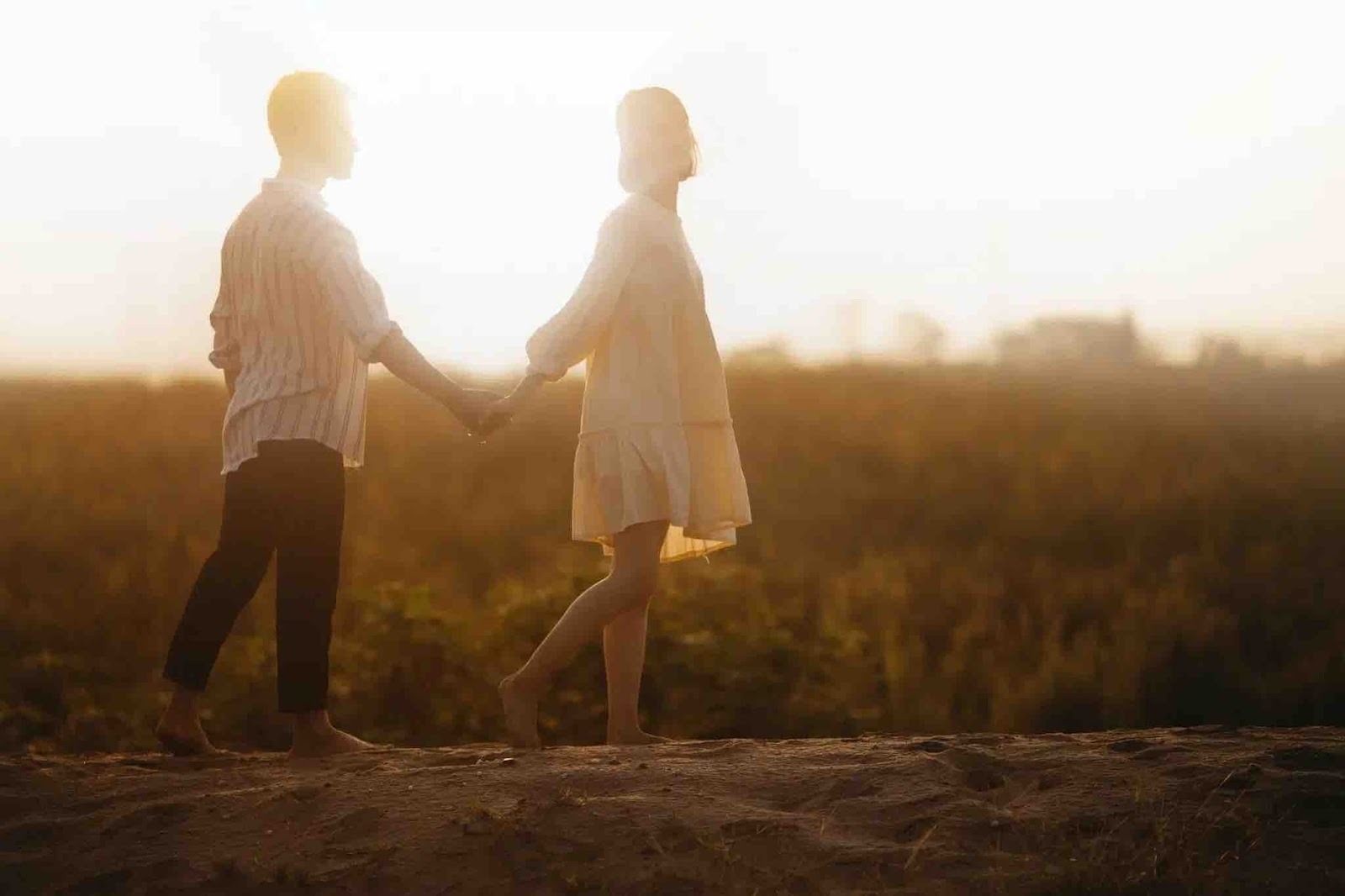 A couple walks hand in hand on a hilltop overlooking a sunlit field, bathed in warm golden light at sunset.