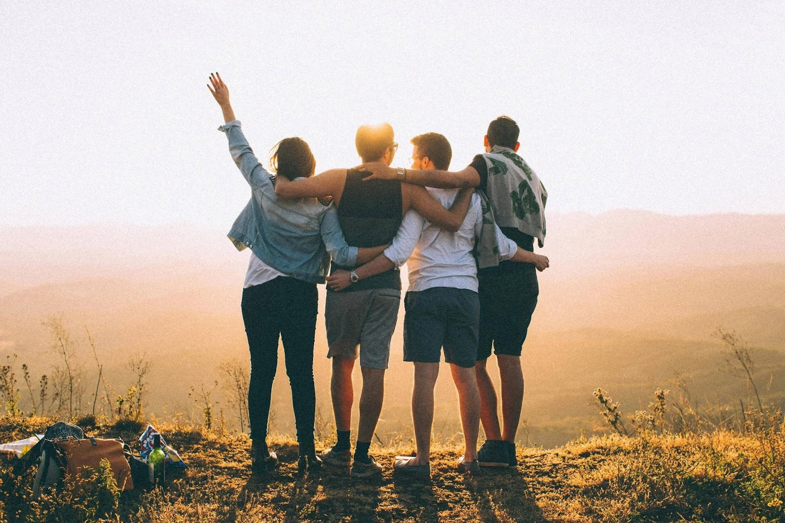 Four friends standing together on a grassy hill at sunset, with their arms around each other's shoulders, looking out toward the horizon.