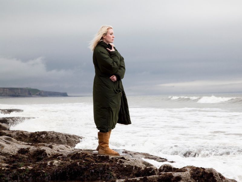 Woman in dark green coat standing on rocky coastline looking out at stormy ocean waves