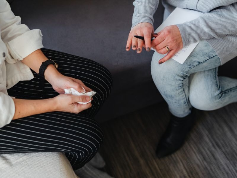 Two people sitting together in a counseling session, one person holding tissues, showing supportive conversation