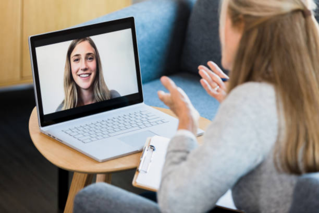 A close-up view of a laptop on a small wooden table showing a smiling therapist during a secure, face-to-face digital counseling session.