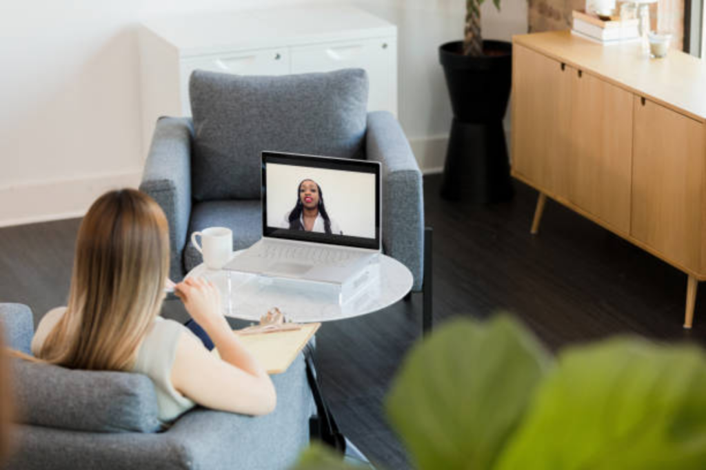 An overhead view of a woman sitting in a gray armchair, looking at a laptop screen where a female therapist is providing telecounseling services.