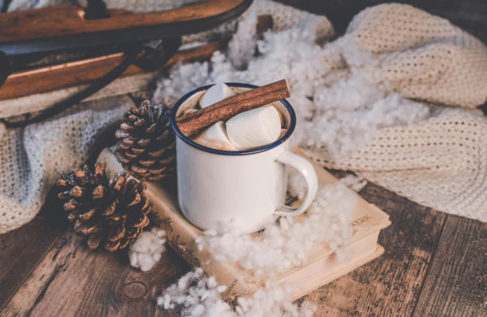 A cozy holiday scene featuring a white mug filled with hot cocoa and large marshmallows, garnished with a cinnamon stick, resting on a vintage book.