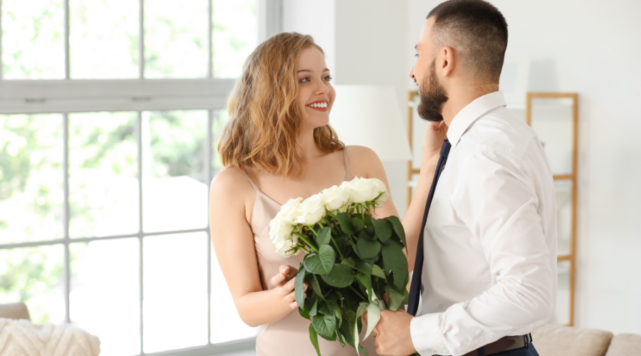 A smiling woman in a beige slip dress receives a bouquet of white roses from a man in a white dress shirt and navy tie, standing in a bright, softly lit living room