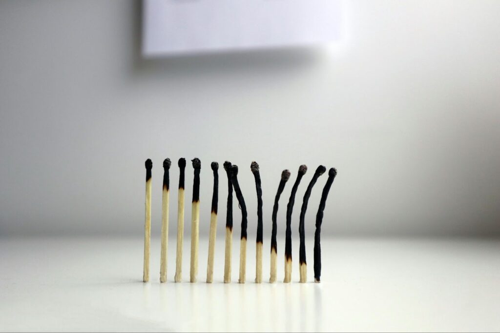A row of matches standing upright on a white surface. The matches on the far left are mostly intact, but as the line moves to the right, the matches become increasingly charred, blackened, and bent downward, visually illustrating the stages of burnout.