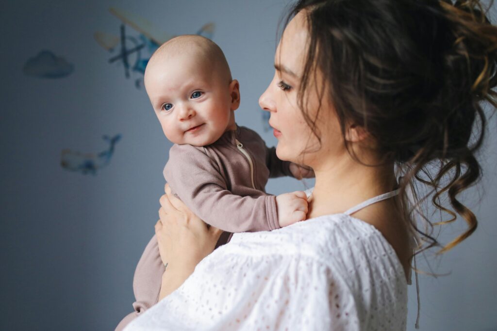 Mother holding a baby close to her shoulder in a softly lit room, the infant looking outward with a gentle smile while the mother gazes at them with a calm, attentive expression.