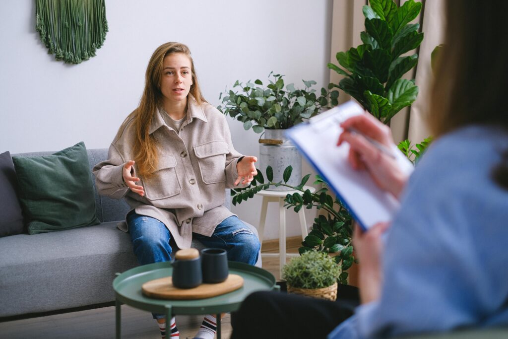 A young woman gestures expressively while speaking to a therapist or counselor who is taking notes on a clipboard.