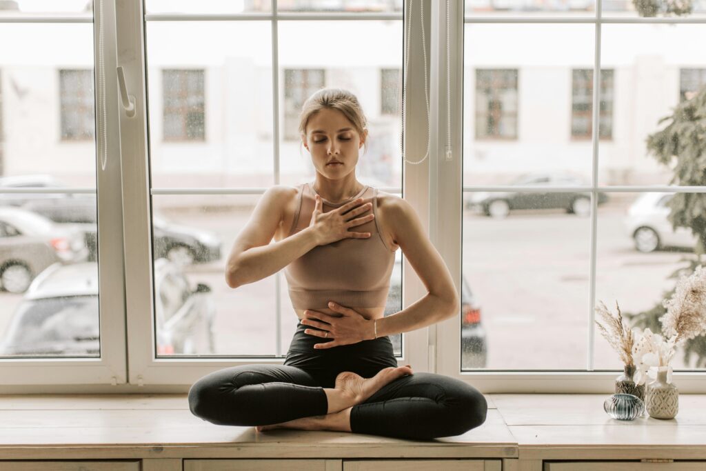 A woman sits cross-legged on a windowsill with eyes closed, one hand on her chest and one on her stomach, practicing breathwork or a somatic grounding exercise for nervous system regulation.