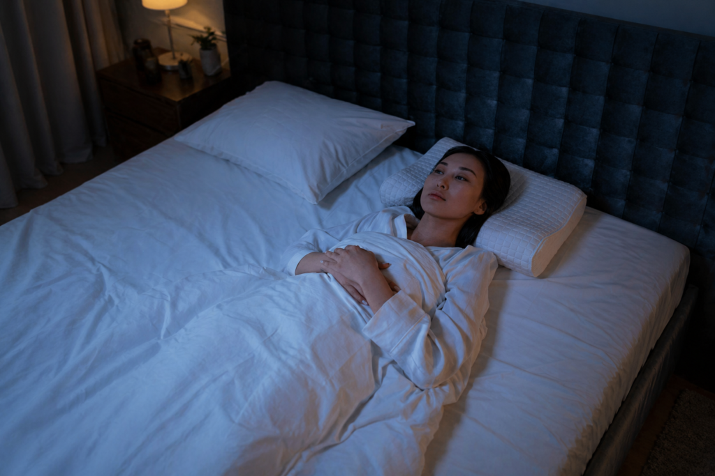 Person lying awake in bed at night, resting on a pillow with hands folded over their chest, softly lit by a bedside lamp, capturing a quiet, restless moment often associated with Sunday night anxiety.