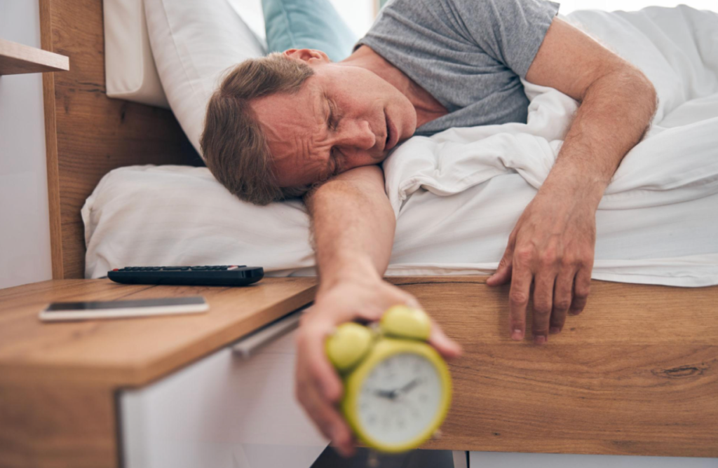 Alt Text: Man lying in bed reaching for an alarm clock, showing symptoms of depression such as fatigue, low energy, and difficulty waking up