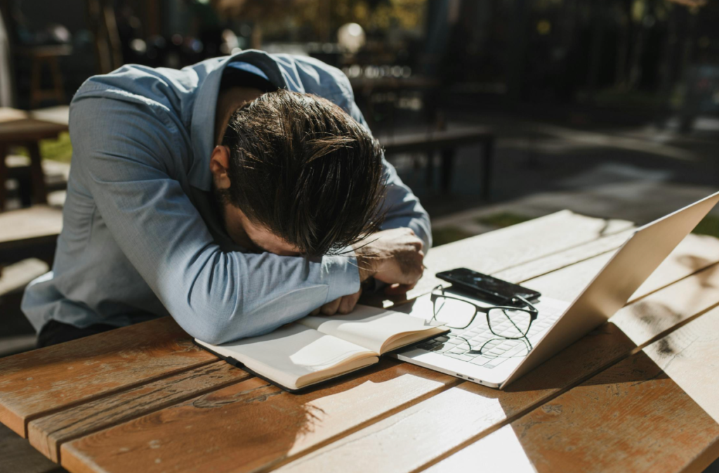  Person resting their head on folded arms at an outdoor table beside a laptop and notebook, appearing mentally drained in bright daylight, capturing the feeling of burnout and exhaustion.
