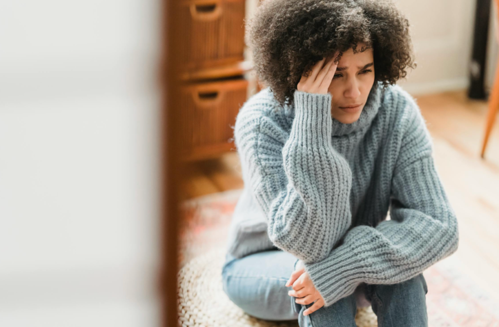 Person sitting on the floor indoors with knees pulled in, resting their head against their hand with a concerned expression, capturing feelings of stress, overwhelm, and emotional fatigue.
