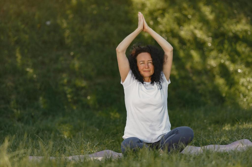 Person sitting cross-legged outdoors on a yoga mat, arms raised overhead with hands together, practicing a calming stretch in a peaceful green setting, representing relaxation and stress relief.
