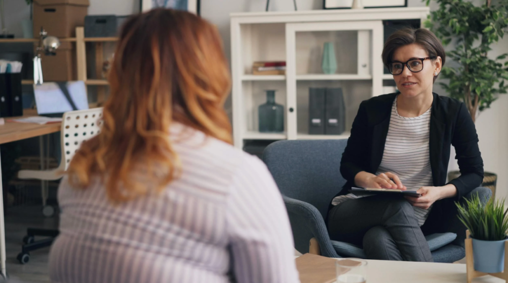  Two people sitting across from each other in a professional office setting, one taking notes while listening attentively, representing a supportive conversation around mental health or stress management.