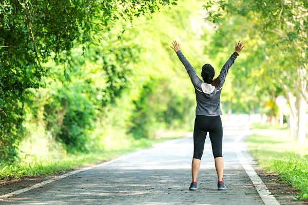 A woman standing outside in a lush green park with her arms raised, finding relief and renewed energy.