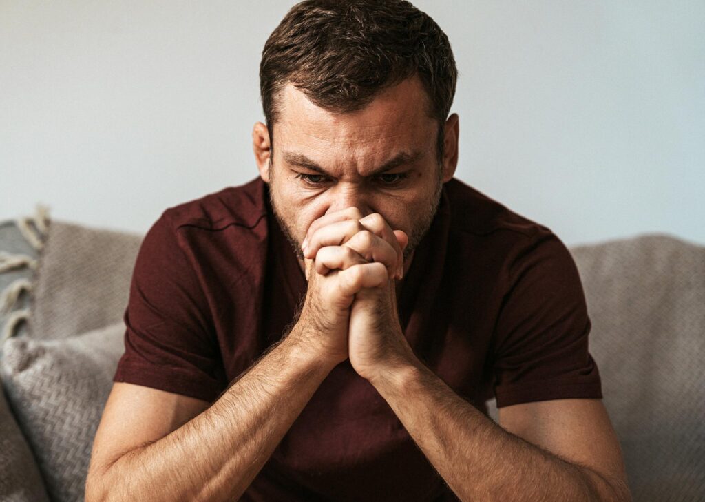 Alt text: Man sitting forward with hands clasped and tense posture, showing signs of depression in men such as stress, rumination, and emotional overwhelm