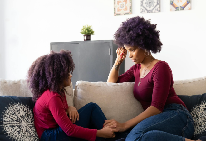 Parent sitting closely with their child on a couch, holding their hands and listening attentively, creating a calm and supportive moment of connection and reassurance during an emotional conversation.