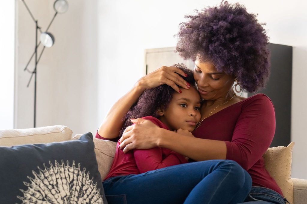 A supportive parent gently hugging a pensive young child on a couch, representing the crucial emotional support needed when navigating youth mental health and difficult conversations.