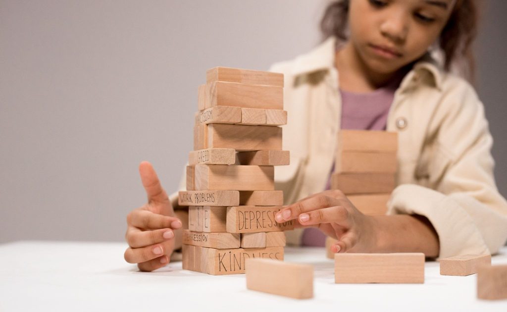 A young child carefully removes a wooden block labeled 'DEPRESSION' from a precarious tower, symbolizing the delicate balance of youth mental health and the importance of parental support.